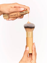 Makeup brush with a gold handle and gray bristle head, held by a hand against a white background.