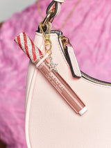Lip gloss attached to a pink handbag. The gloss has red and white striped cap with rhinestones and a charm. Text reads: Tarte maracuja juicy lip plump. Background is soft pink fabric.