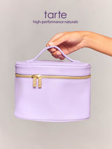 A lavender makeup bag with a handle is held by a hand, showcasing a zippered closure with two gold zippers labeled Tarte, against a plain, light gray background.